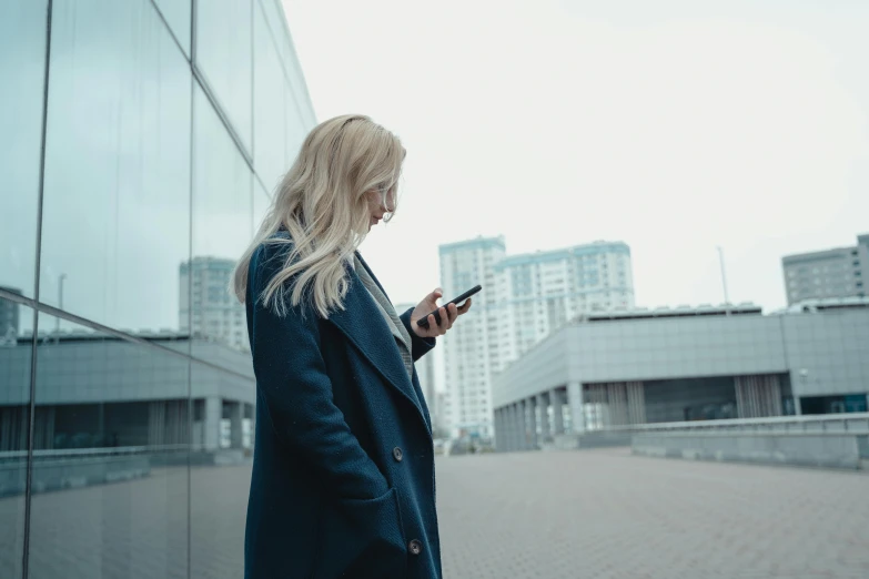 a woman standing in front of a building looking at her cell phone, pexels contest winner, a girl with blonde hair, wearing a long coat, female in office dress, mid 2 0's female