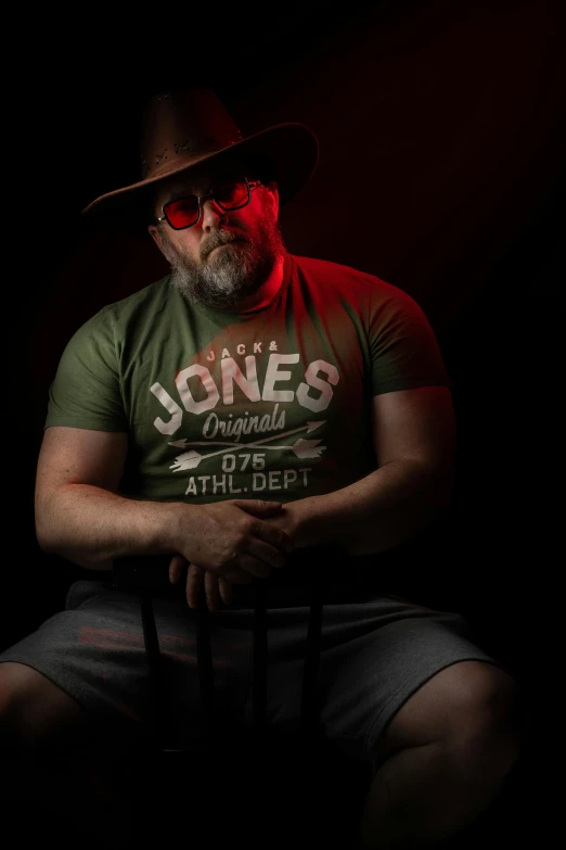a man sitting on a chair in a dark room, a character portrait, inspired by James Dickson Innes, unsplash, wearing sunglasses and a hat, gabe newell as a superman, red and green lighting, portrait of rugged male ranger