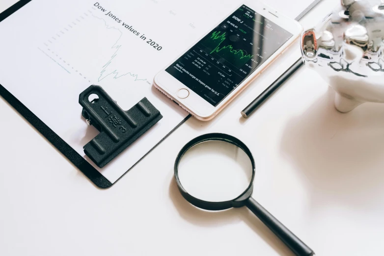 a cell phone sitting on top of a clipboard next to a magnifying glass, a picture, by Adam Marczyński, trending on pexels, displaying stock charts, on a white table, three fourths view, various posed