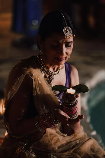 a woman sitting next to a pool holding a lit candle, dressed in a sari, ceremony, lights off, feast