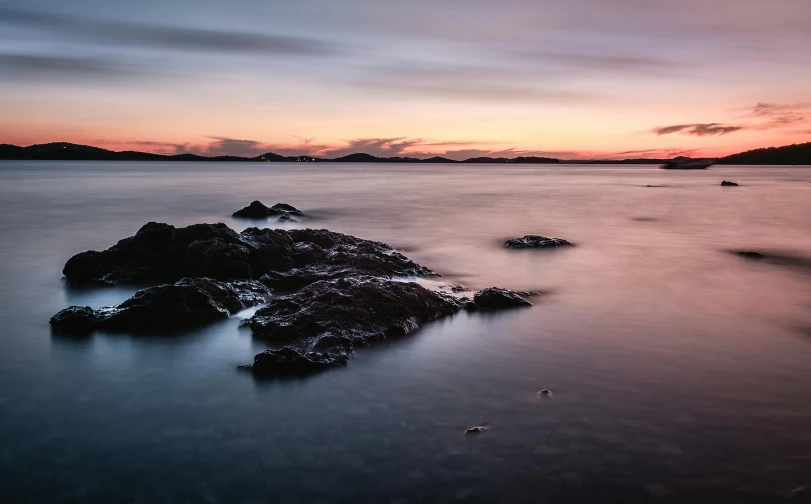 a sunset over a body of water with rocks in the foreground, by Alexander Robertson, pexels contest winner, islands on horizon, grey, manly, subtle colors