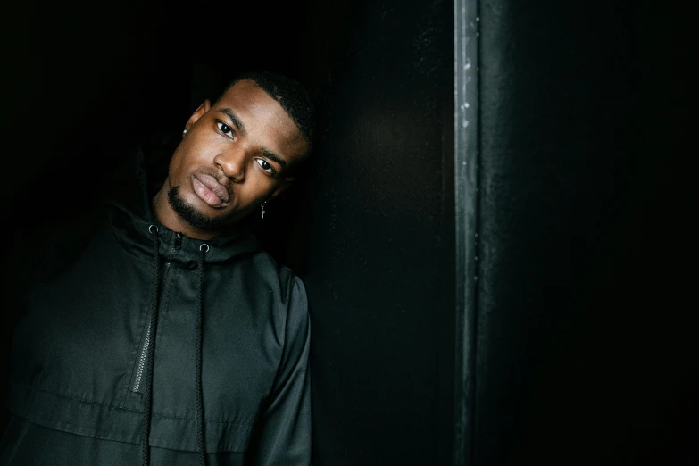 a man in a black jacket leaning against a wall, an album cover, inspired by Theo Constanté, unsplash, maria borges, wearing a black hoodie, looking to camera, concert