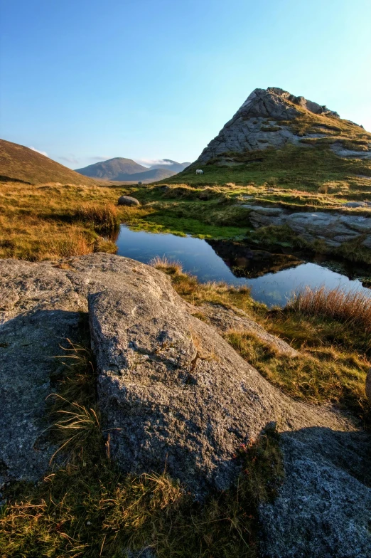 a body of water sitting on top of a grass covered hillside, irish mountains background, sharp spiky rocks, morning golden hour, ponds