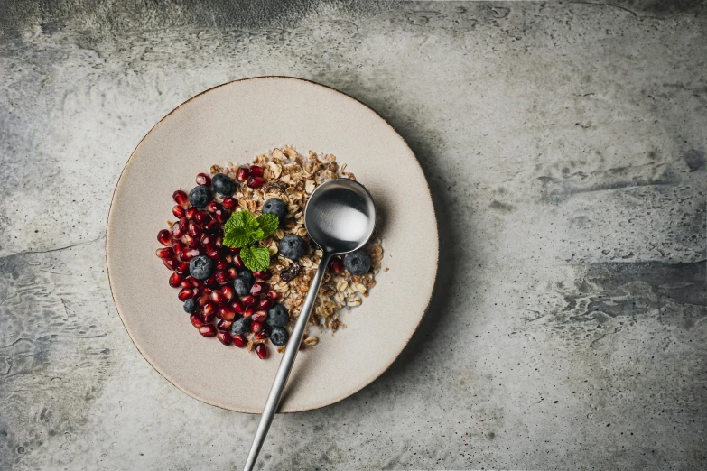 a white plate topped with oatmeal and berries, a still life, trending on pexels, pomegranate, spoon placed, background image, grey