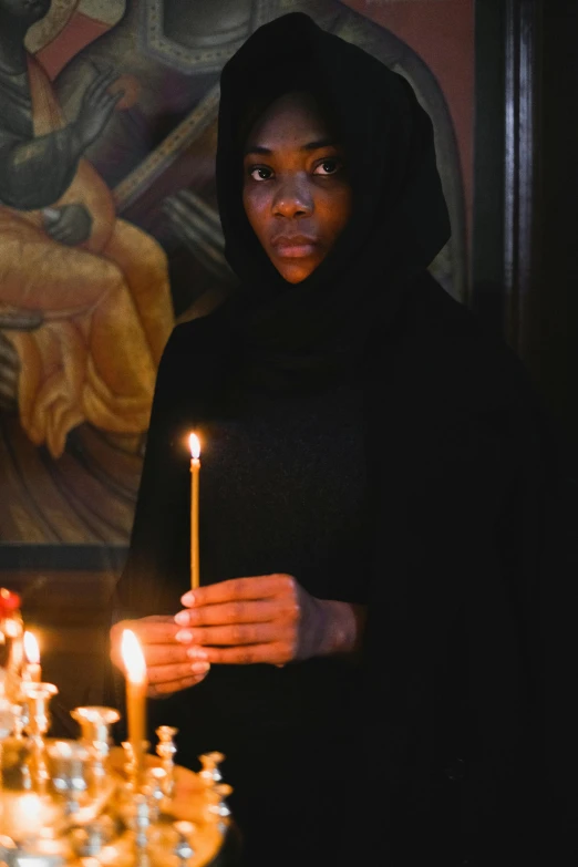 a woman holding a lit candle in front of a painting, black robes, dark skinned, in orthodox church, profile image