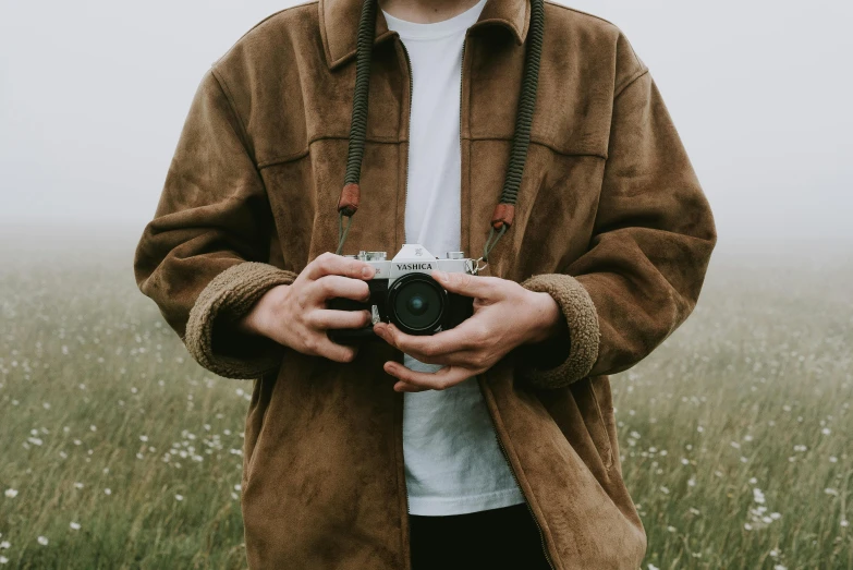 a man standing in a field holding a camera, a picture, pexels contest winner, brown jacket, simple aesthetic, traveling clothes, high details on clothes