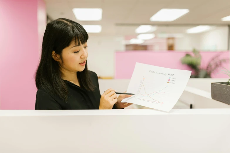 a woman sitting at a desk looking at a piece of paper, a cartoon, pexels contest winner, half asian, graphs, standing straight, nursing