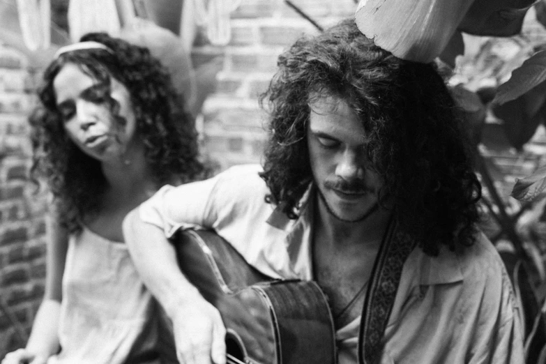 a man playing a guitar next to a woman, by Bruce Davidson, vanitas, long messy curly hair, live performance, bongos, curls on top of his head