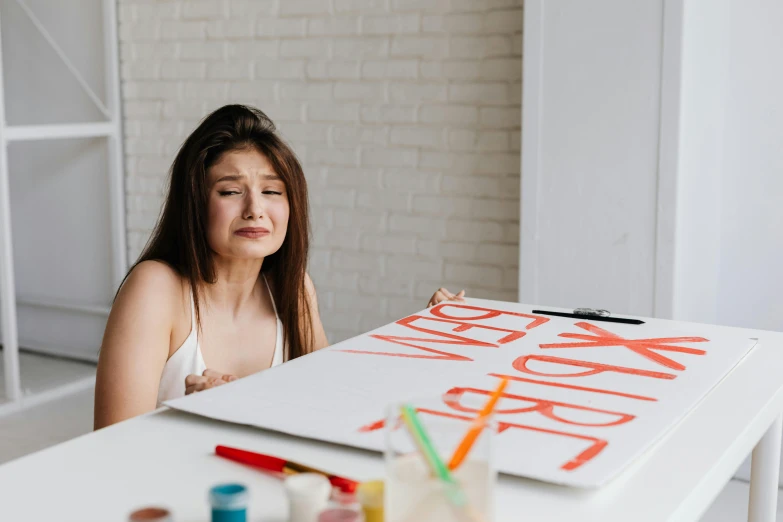 a woman sitting at a table with a sign on it, trending on pexels, action painting, woman crying, paint on canvas, college, on a white table