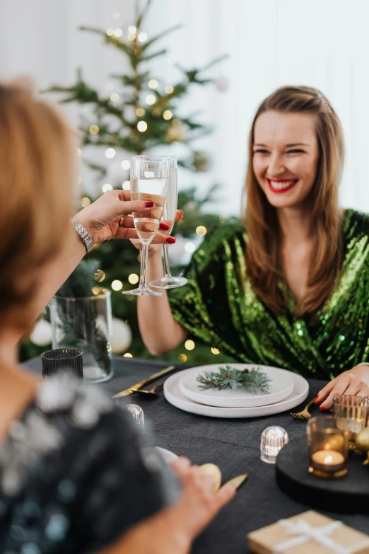 two women sitting at a table with a christmas tree in the background, pexels contest winner, renaissance, cheers, place setting, green sparkles, wearing festive clothing