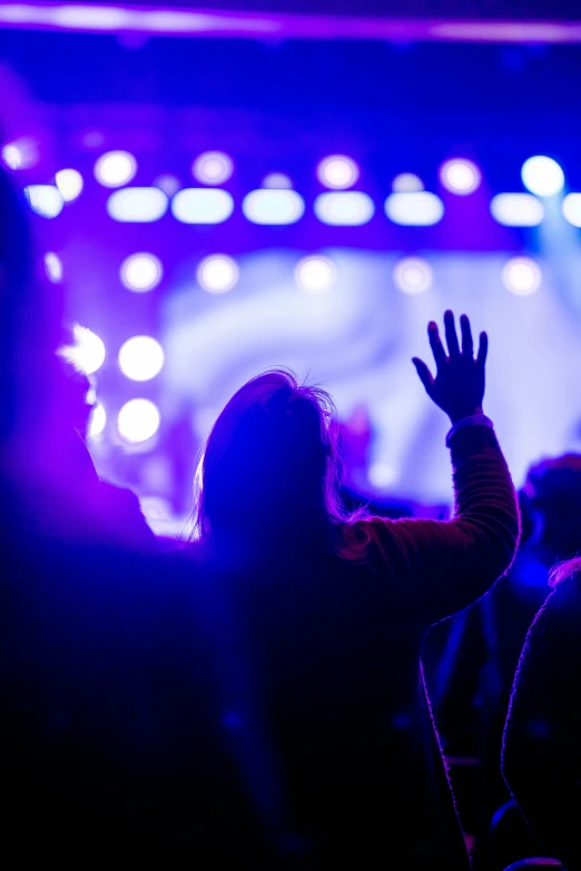 a person raising their hands in front of a stage, pexels, blue and purple lighting, jesus wasted at a party, people walking around, listening to godly music