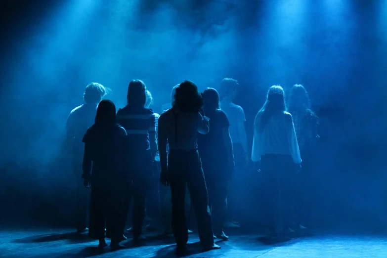 a group of people standing on top of a stage, by Christen Dalsgaard, shutterstock, antipodeans, dramatic blue light, facing away from the camera, theatrical, teenage girl