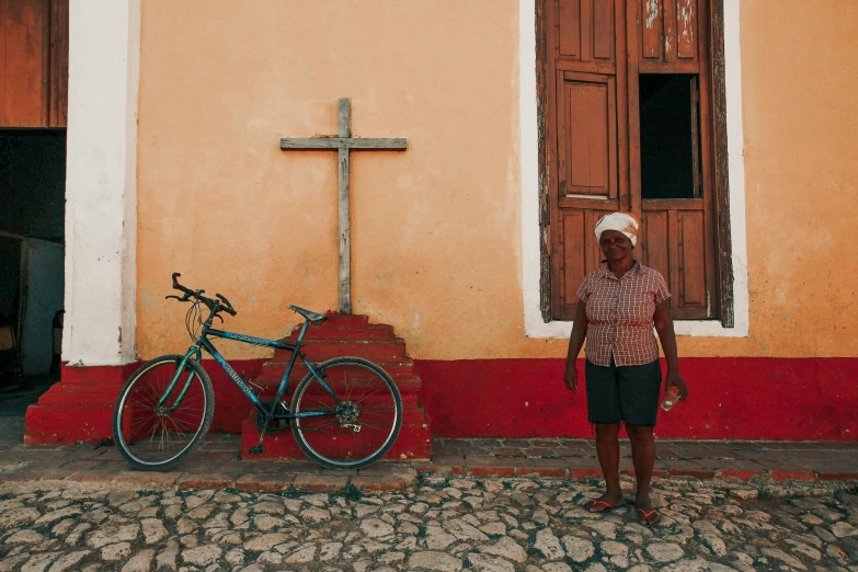 a man standing next to a bike in front of a building, by Elsa Bleda, pexels contest winner, standing in a church, 30-year-old woman from cuba, an old lady with red skin, slide show