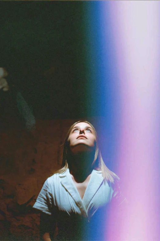 a woman standing in front of a colorful light, unsplash, light and space, grainy movie still, young woman looking up, lightshafts, promotional image