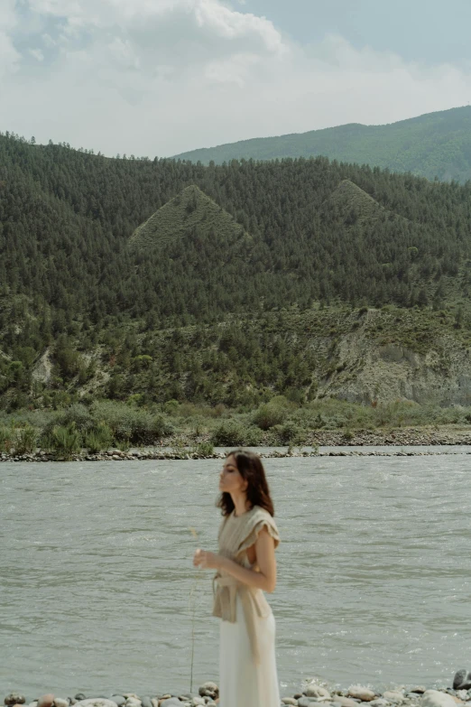 a woman standing next to a body of water, mountains and rivers, movie still frame, azamat khairov, low colour