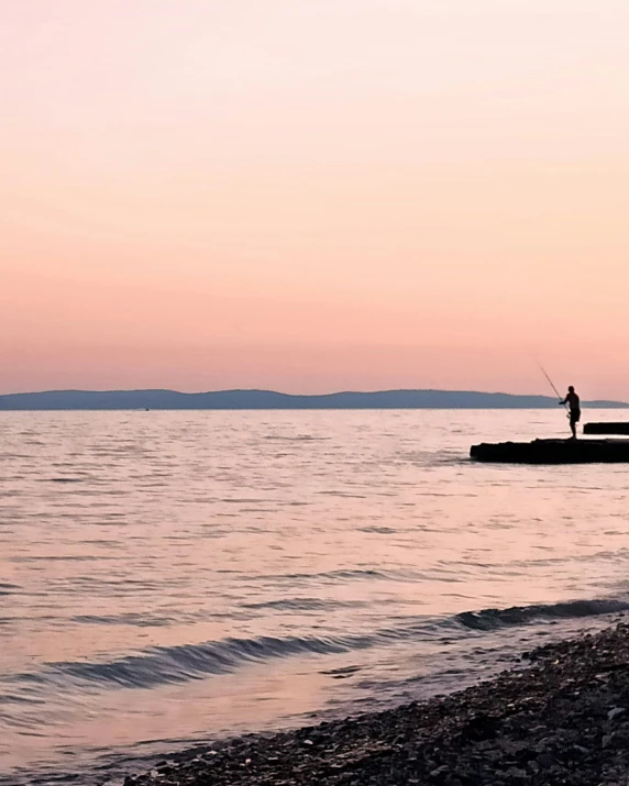 a man standing on top of a beach next to a body of water, a picture, pink skies, fishing, split near the left, lgbtq