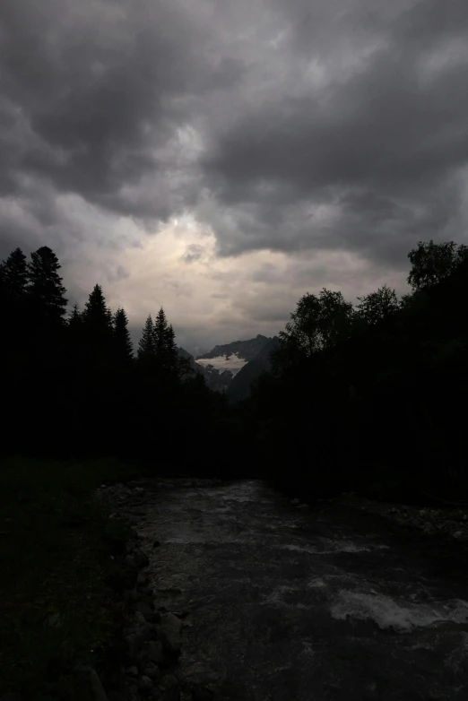 a river running through a forest under a cloudy sky, by Muggur, hurufiyya, gloomy lighting!!!, lightening, in the swiss alps, dark eerie pic