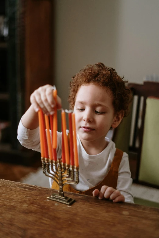 a little boy sitting at a table with a menorah, pexels, instagram post, curls, orange hue, holding a torch