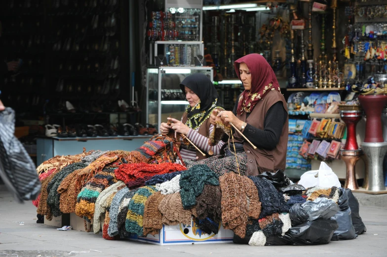 a couple of women sitting next to each other on a street, flickr, hurufiyya, crafts and souvenirs, crochet, youtube thumbnail, lots of shops