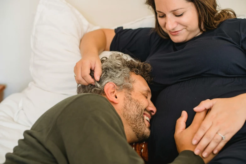 a woman sitting next to a man on a bed, by Carey Morris, pexels contest winner, happening, pregnant belly, head bent back in laughter, profile image, coloured