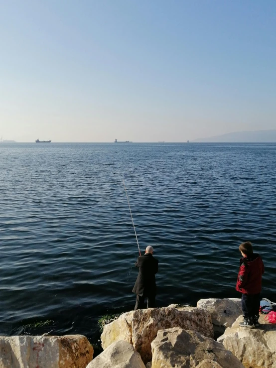 a couple of people standing on top of a rock next to a body of water, fishing, at the sea, split near the left, floating power cables