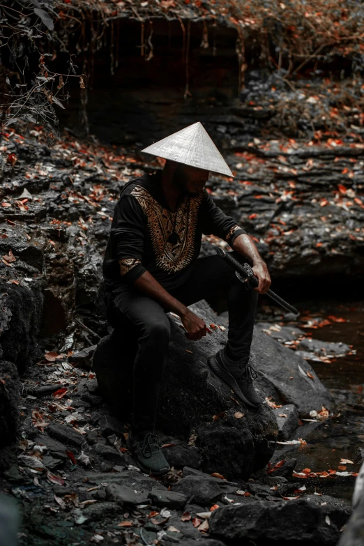 a man sitting on top of a rock next to a river, an album cover, inspired by Ma Quan, pexels contest winner, afrofuturism, black pointed hat, samurai outfit, malaysia jungle, camo