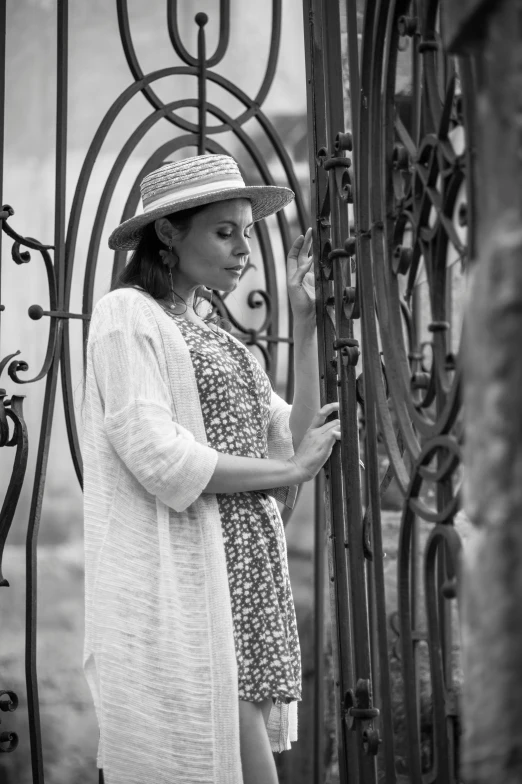 a black and white photo of a woman standing in front of a gate, a black and white photo, pexels contest winner, straw hat and overcoat, wrought iron, photoshooting, dressed in a beautiful white