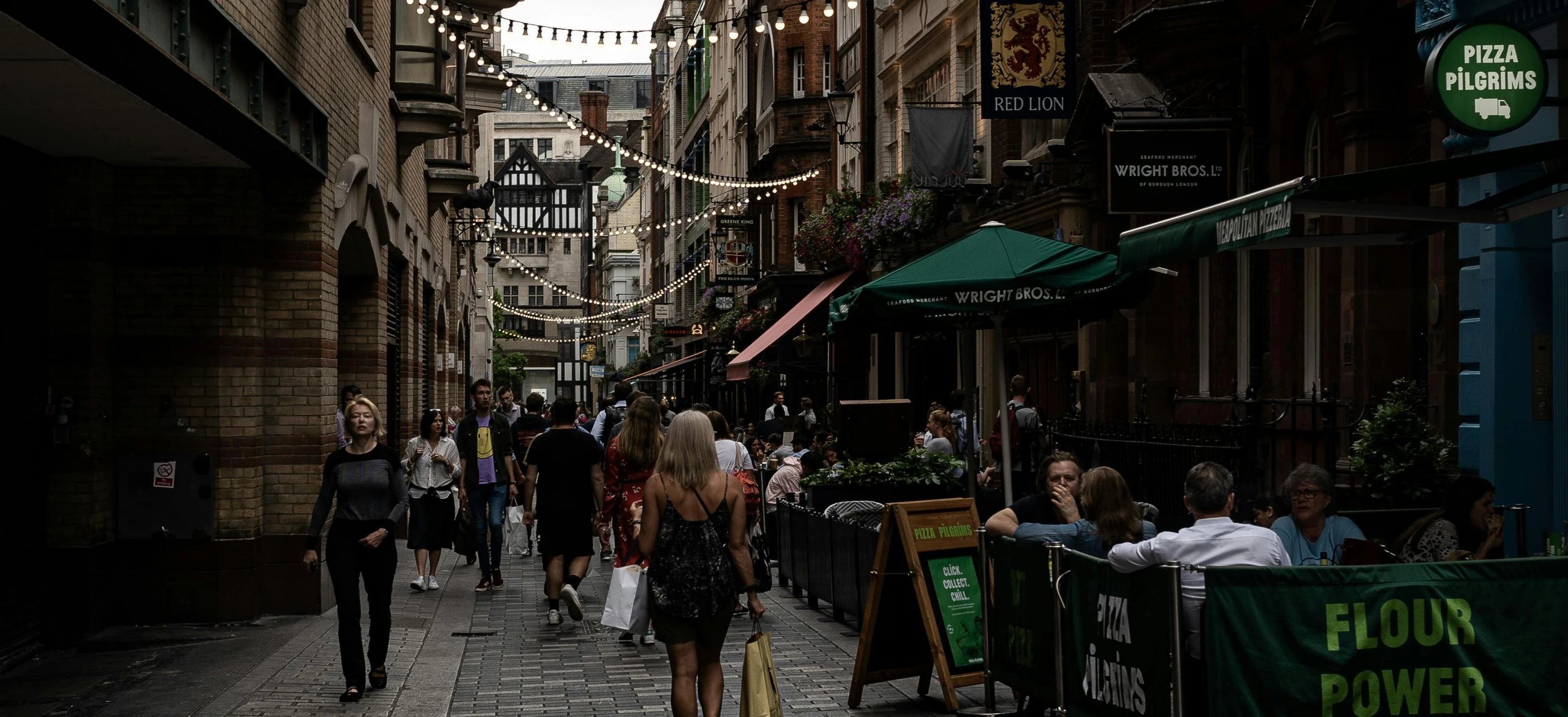 a group of people walking down a street next to tall buildings, pub, a busy arcade, unsplash photography, summer lighting