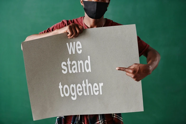 a man holding a sign that says we stand together, trending on pexels, avatar image, background image, high school, masking