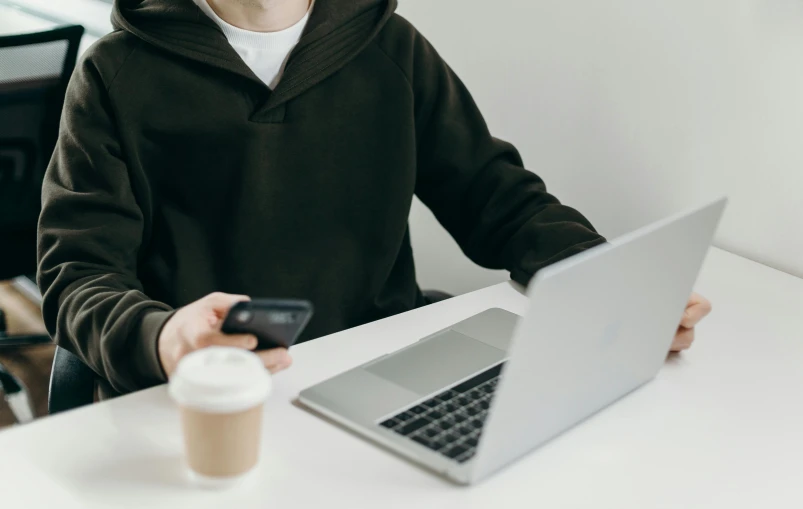 a man sitting at a table with a laptop and a cup of coffee, by Carey Morris, trending on pexels, wearing a black hoodie, minimalist logo without text, phone in hand, link from zelda using computer