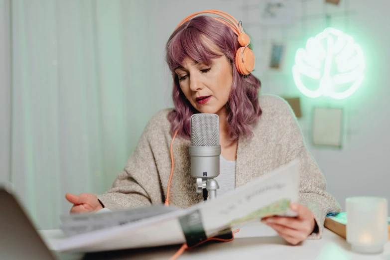 a woman sitting at a table in front of a laptop, an album cover, trending on pexels, sitting in front of a microphone, green and purple studio lighting, reading a newspaper, wearing modern headphone