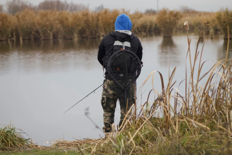 a person standing next to a body of water, people angling at the edge, with a backpack, helmond, baggy clothing and hat