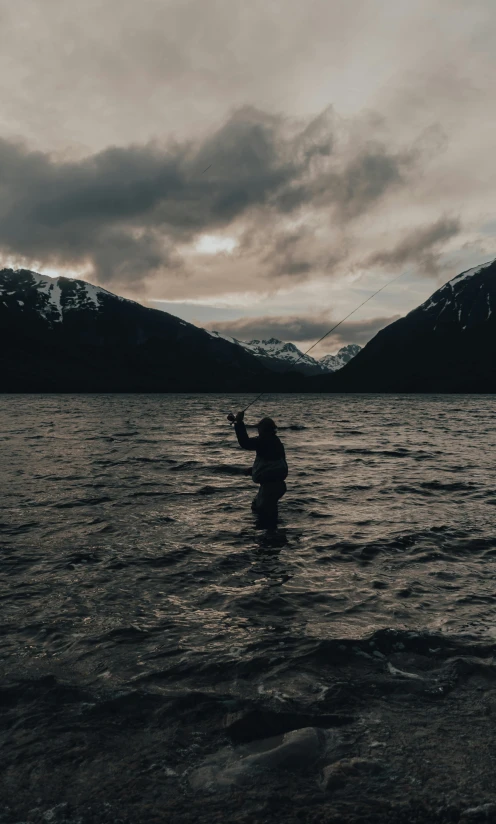 a person in a body of water with mountains in the background, fishing, it's getting dark, landscape photo, waving