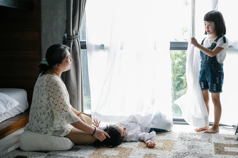 a woman sitting on the floor next to a little girl, by Lucia Peka, pexels contest winner, curtains, wearing white pajamas, husband wife and son, space seen outside from a window