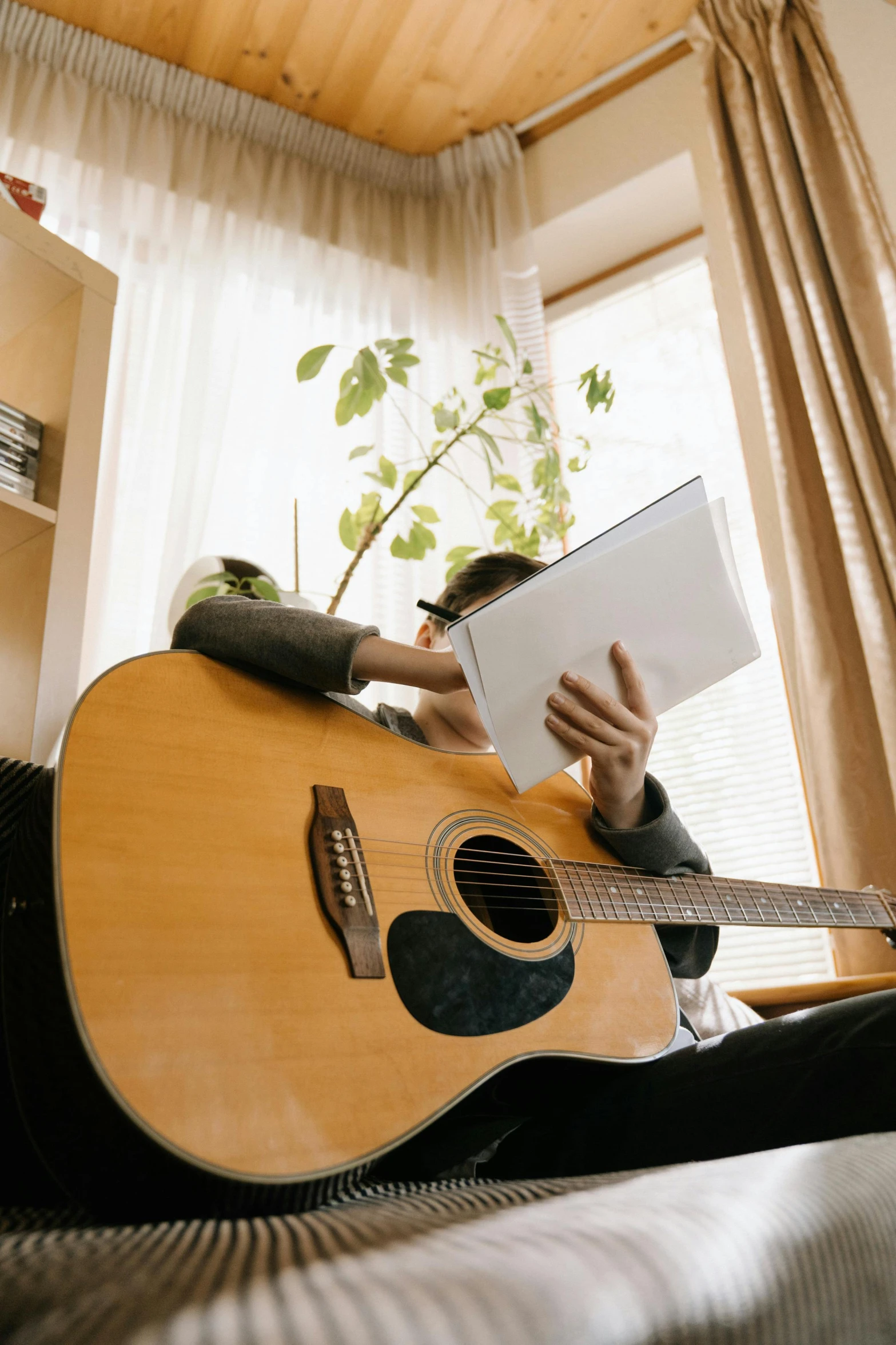 a person laying on a bed with a guitar, holding a book, window open, full device, acoustic guitar