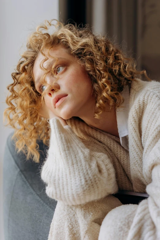 a woman sitting on top of a chair next to a window, inspired by Louisa Matthíasdóttir, trending on pexels, renaissance, curly hair, wearing casual sweater, thoughtful pose, on a couch