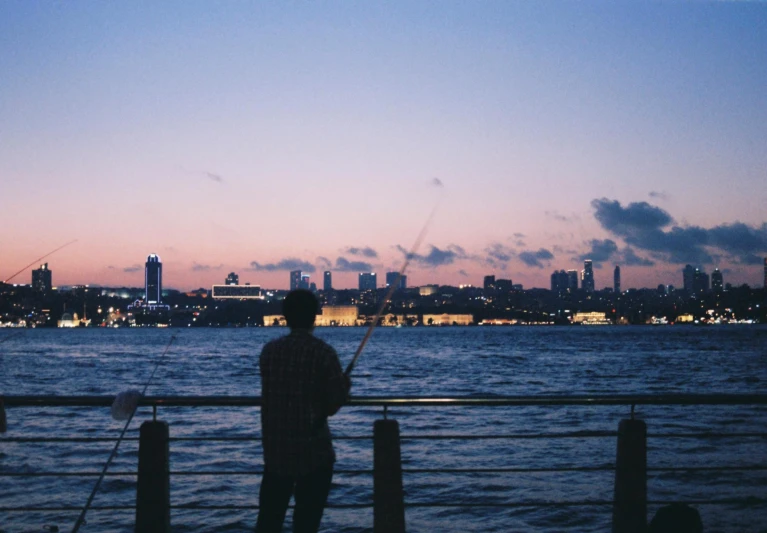 a man standing on top of a pier next to a body of water, inspired by Elsa Bleda, pexels contest winner, happening, brooklyn background, twilight skyline, fishing, overlooking the ocean