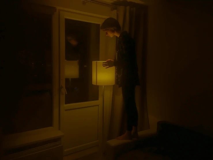 a person standing next to a lamp in a dark room, near a window, lit. 'the cube', cinematic pastel lighting, laura watson