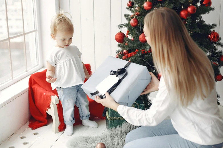 a woman holding a baby in front of a christmas tree, pexels contest winner, holding gift, grey, blank, indoor scene