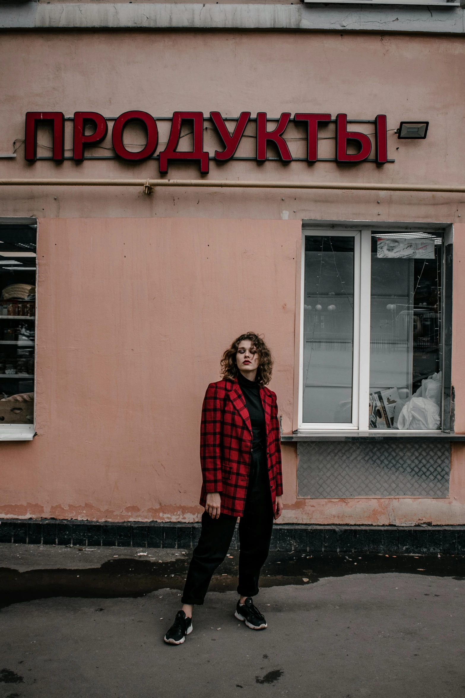 a woman standing in front of a pink building, russian style, black and red jacket, non-binary, stood in a supermarket