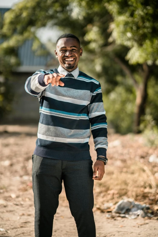 a man standing on a dirt road pointing at the camera, wearing stripe shirt, teaching, wearing a sweater, unmistakably kenyan