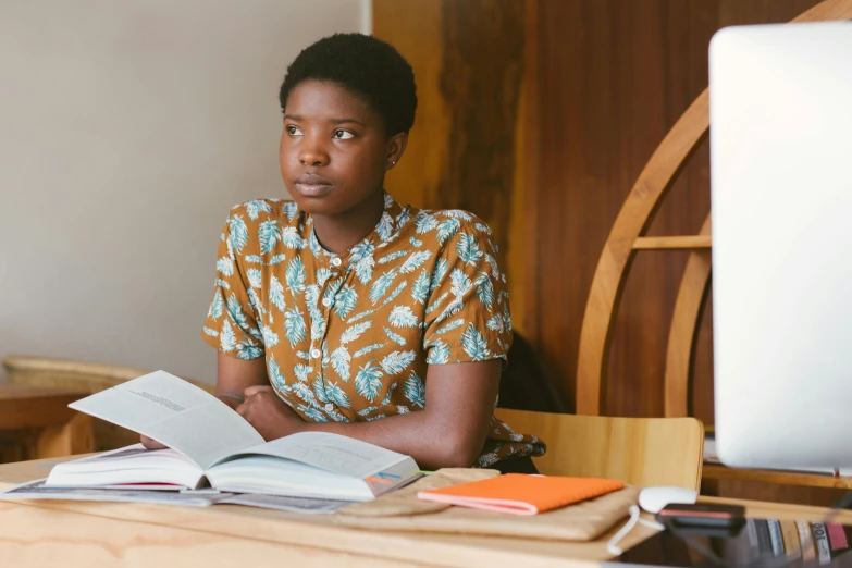 a woman sitting at a desk in front of a computer, a portrait, by Lily Delissa Joseph, pexels, hurufiyya, lupita nyong'o, with one vintage book on a table, looking serious, wearing yellow floral blouse