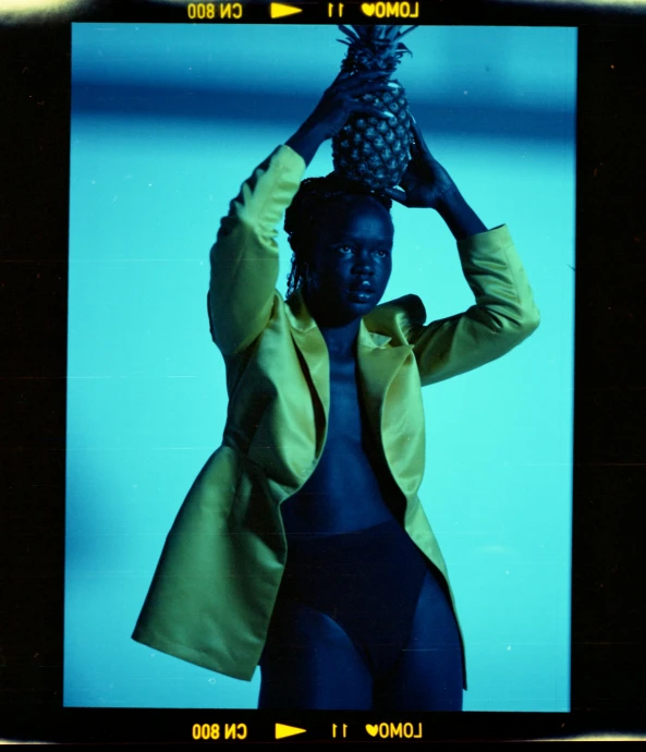 a woman holding a pineapple on top of her head, unsplash, afrofuturism, blue skintight closed longcoat, fujicolor with flash, trench coat and suit, black and yellow