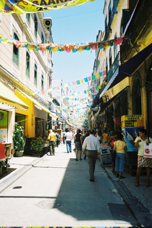 a group of people walking down a street next to tall buildings, streets of salvador, yellow awning, in 2 0 0 2, old shops