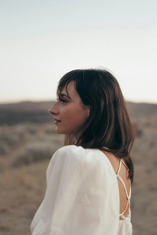 a woman in a white dress standing in the desert, an album cover, inspired by Zoë Mozert, head and shoulders view, side portrait rugged girl, 2019 trending photo, brown hair and bangs