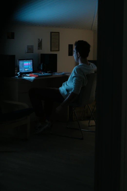 a person sitting at a desk in a dark room, an album cover, computer art, student, movie filmstill, indoor picture, programming