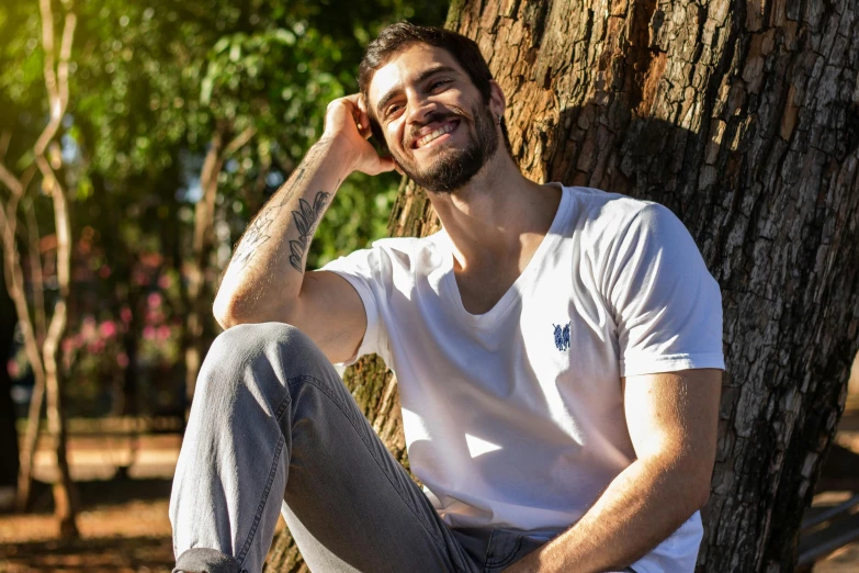 a man sitting next to a tree talking on a cell phone, plain white tshirt, brazilan supermodel, while smiling for a photograph, avatar image