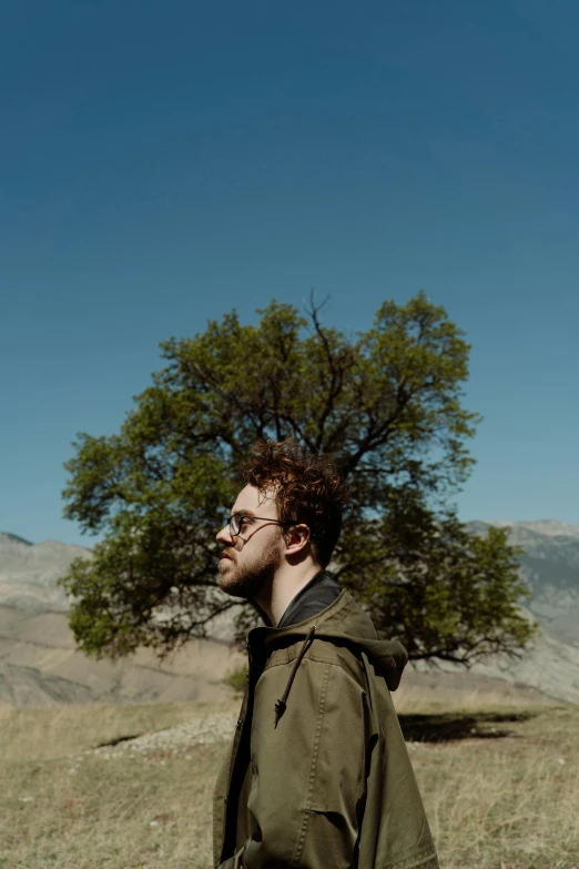 a man standing in a field with a tree in the background, an album cover, unsplash, seth aaron rogen, mountains, ((trees)), profile portrait