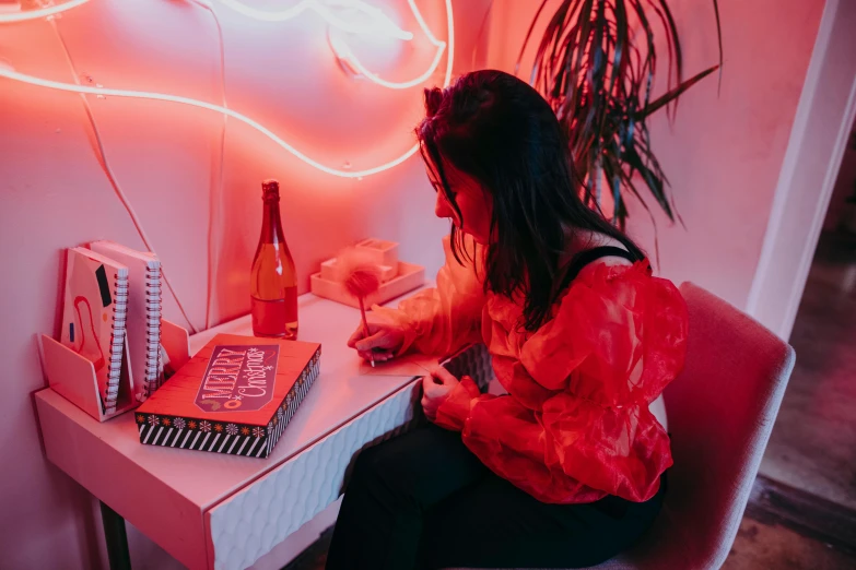 a woman sitting at a table in front of a neon sign, by Sydney Carline, pexels contest winner, red ballpoint pen, writing in journal, the walls are pink, red orange lighting