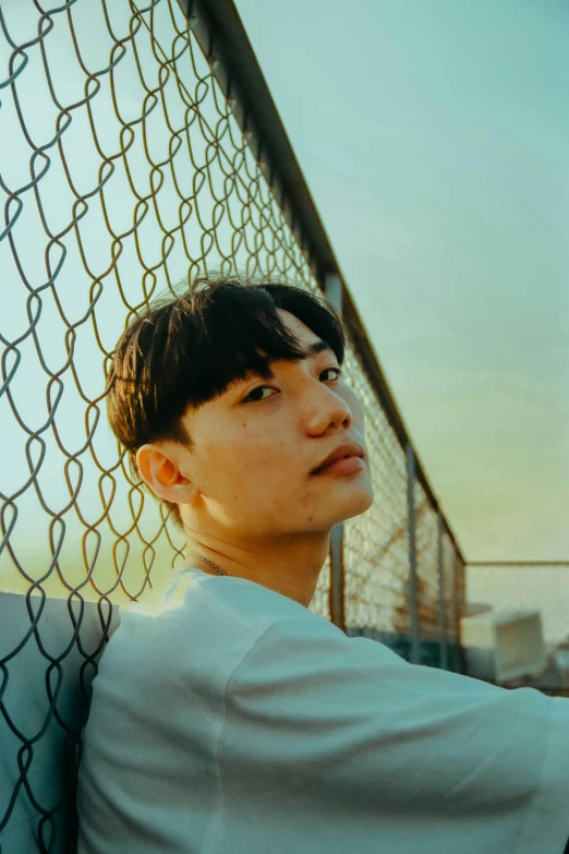 a man leaning against a chain link fence, an album cover, inspired by jeonseok lee, unsplash, realism, young cute wan asian face, sun down, kai carpenter, wrinkly